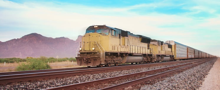 A cargo train passing from the right side of the screen with mountains in the background